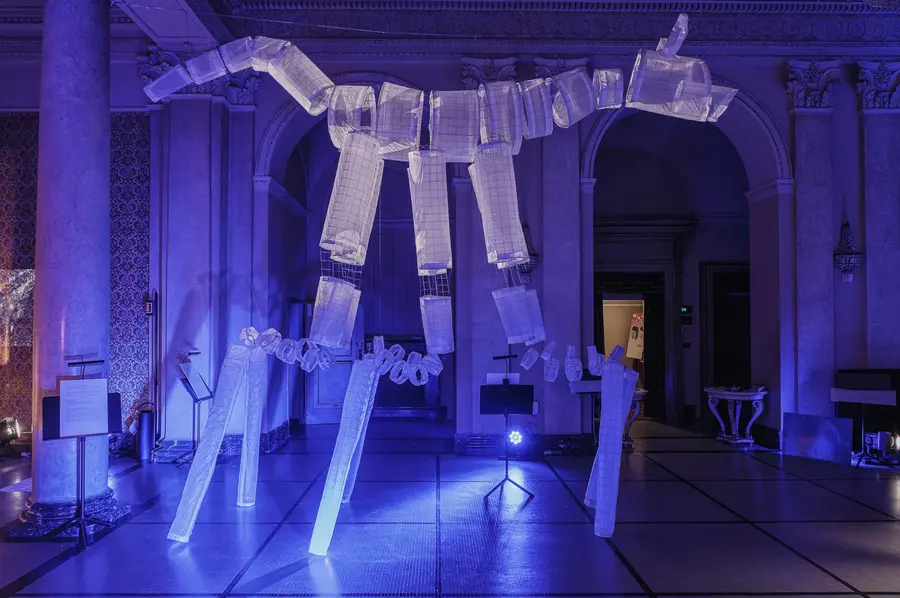 Figura gigante di carta e rete sospesa tra le colonne di una sala, in luce blu